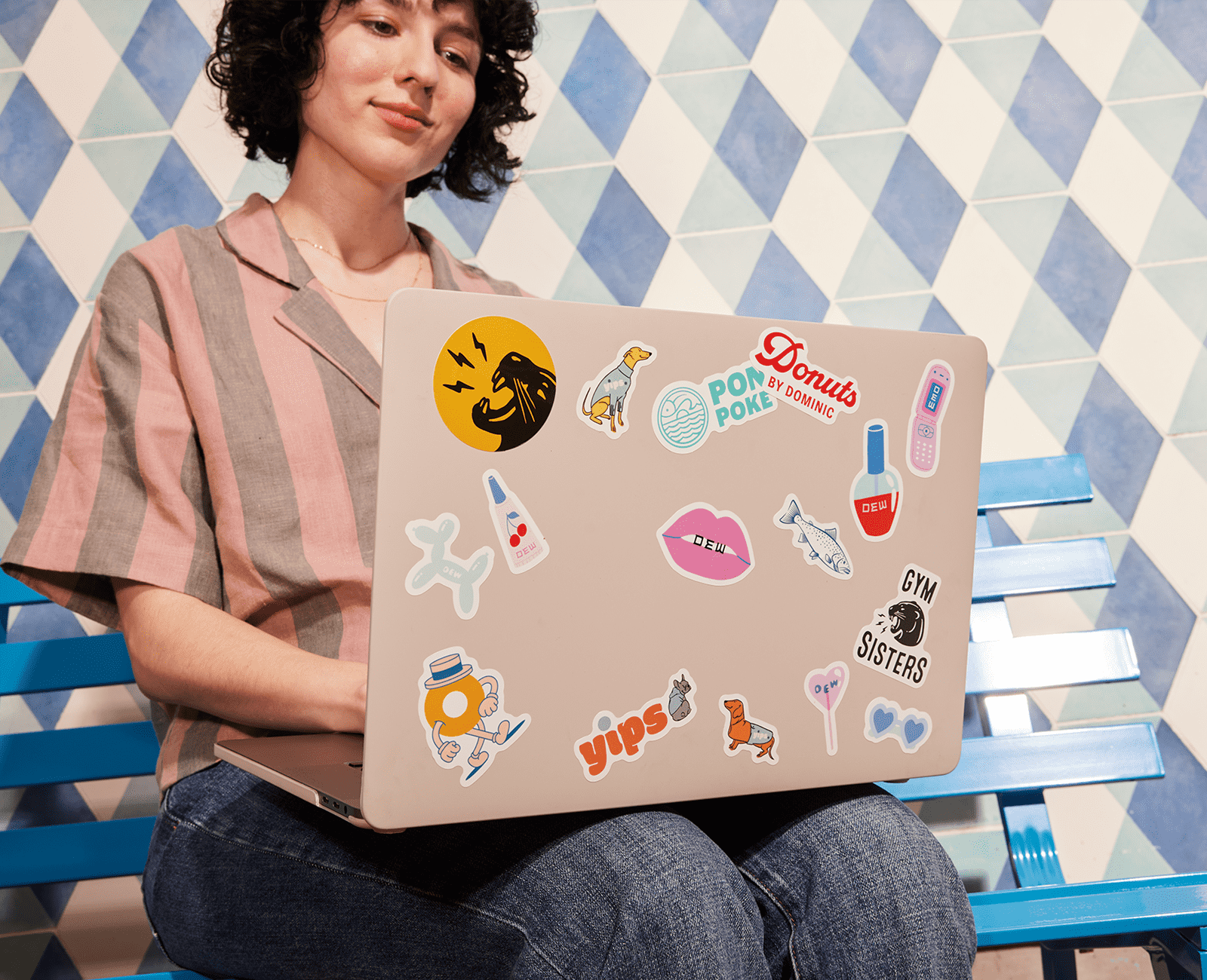 A woman sitting in front of a wall with a white and blue diamond design and using a laptop with stickers on the lid