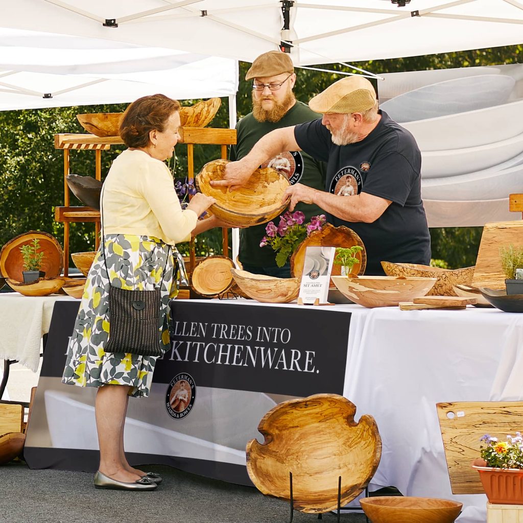 Two vendors at a craft market demonstrate handcrafted wooden kitchenware to a customer at an outdoor booth