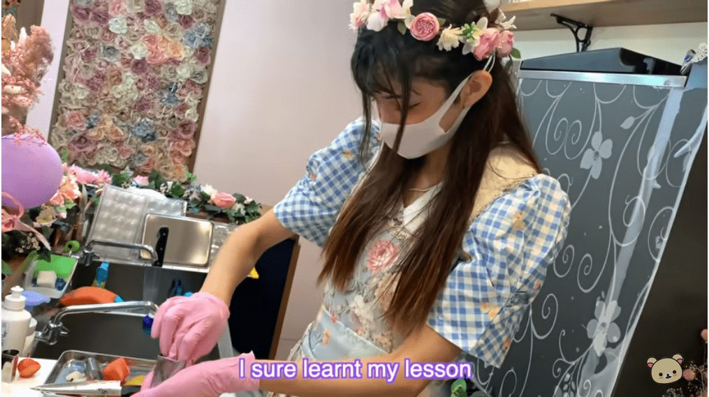  A lady dressed in blue plaid dress and floral apron is seen preparing skewered fruits