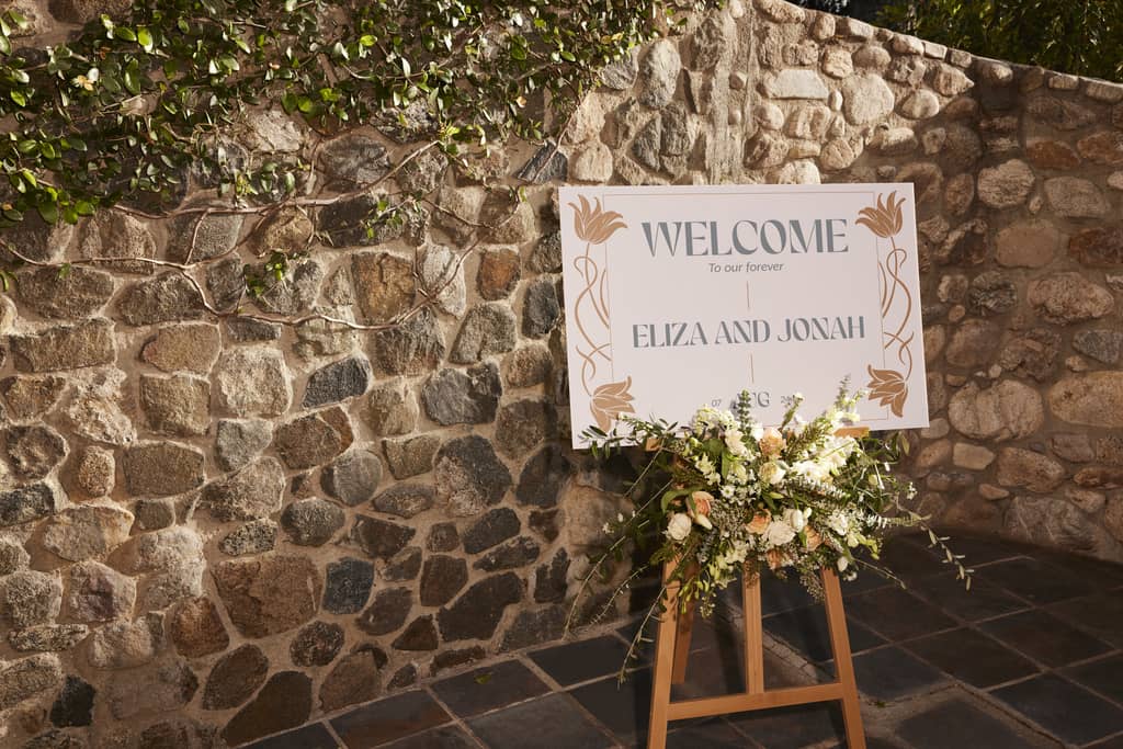 a customized wedding sign in front of a stone wall, with a floral bouquet at the base