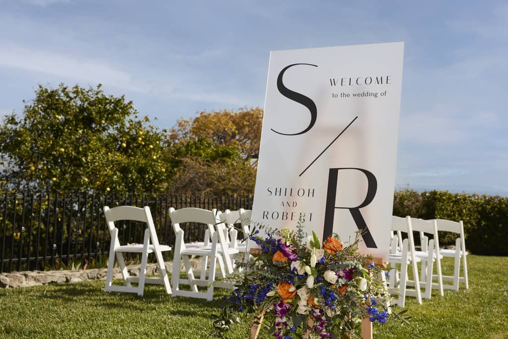 a large wedding sign places outside, above a bouquet of flowers and in front of the seating arrangement