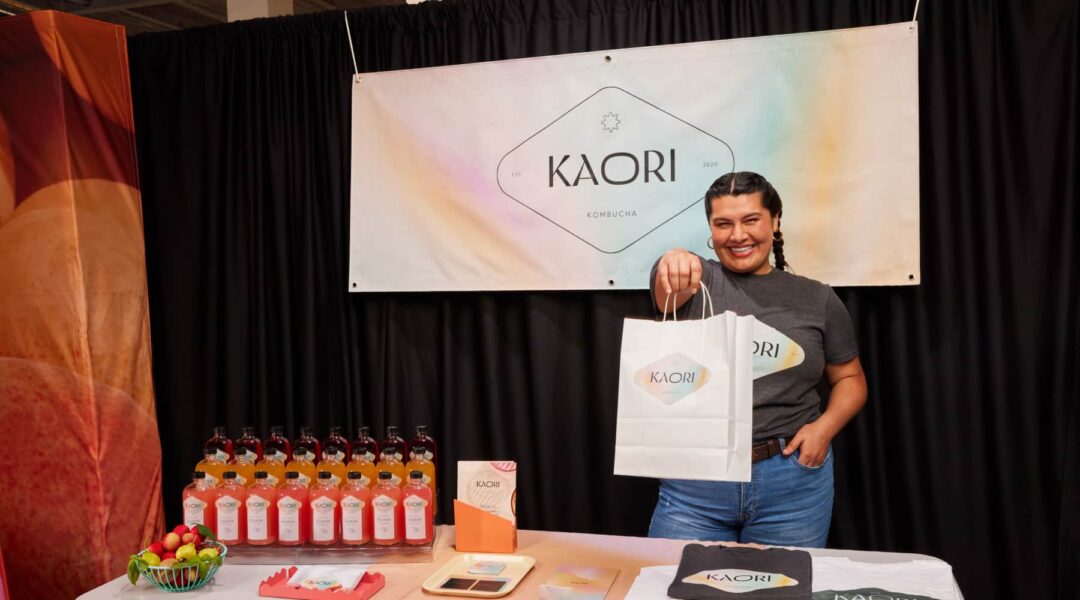 Small business owner at a fair stand holding a custom paper bag with her logo, colorful products on the table and a branded community banner in the background.