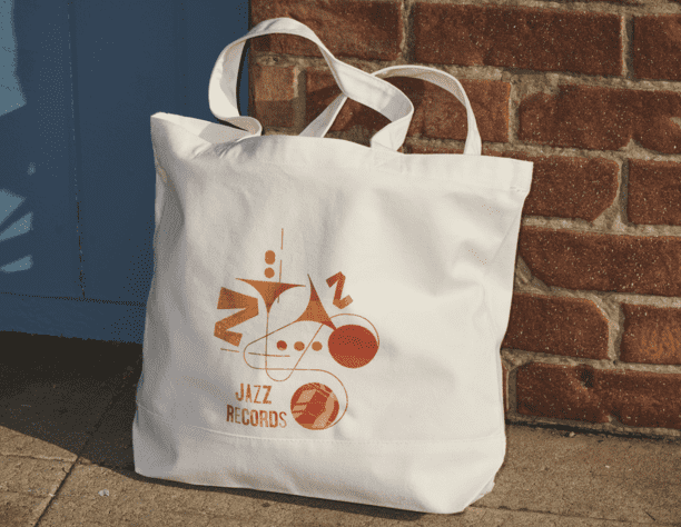 A branded tote sits on the floor against a brick wall. The tote features a branding design with a jazzy orange and yellow interpretation of musical instruments and notes.