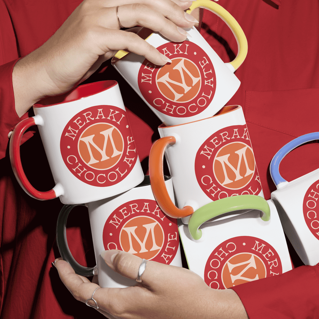 Chest-level shot of woman in red long-sleeved shirt holding several custom mugs branded with an orange &ldquo;M&rdquo; chocolatier logo and featuring mug handles of different colors