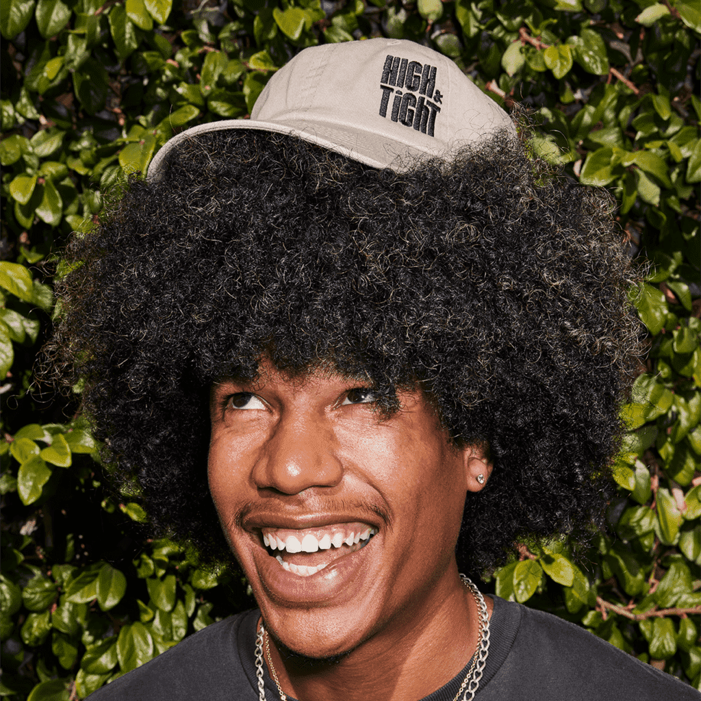 Joyful man with tall curly hair, playfully poses with an embroidered brand hat balanced on his hair.