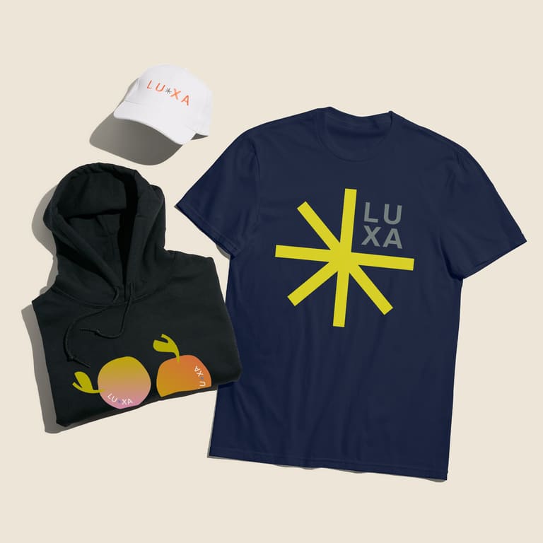 A flatlay of brand merch, featuring a white branded cap, hoodie and a navy blue branded crewneck T-shirt with a brand logo placed at the front