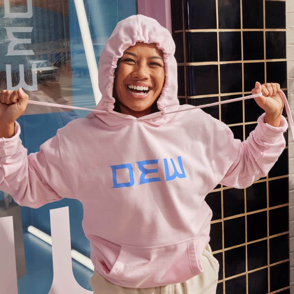 Young woman smiles brightly at camera while wearing a cozy, pink branded hoodie with the hood up.
