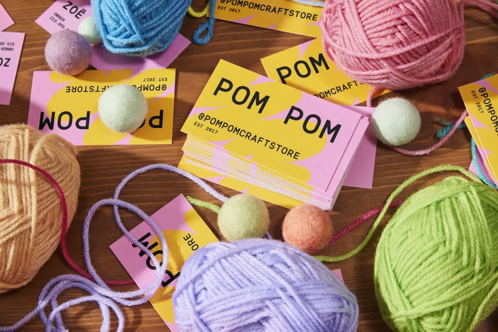 Bright pink and yellow business cards with bold simple black text “Pom Pom” and “@pompomcraftstore” scattered across a wooden table with colorful balls of yarn.