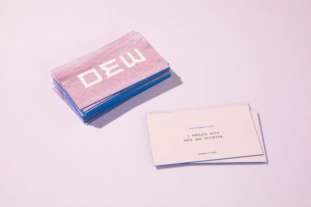 Stack of blue edged pink business cards with large white “DEW” text on one side and small simple black text “I radiate with hope and optimism on the other.