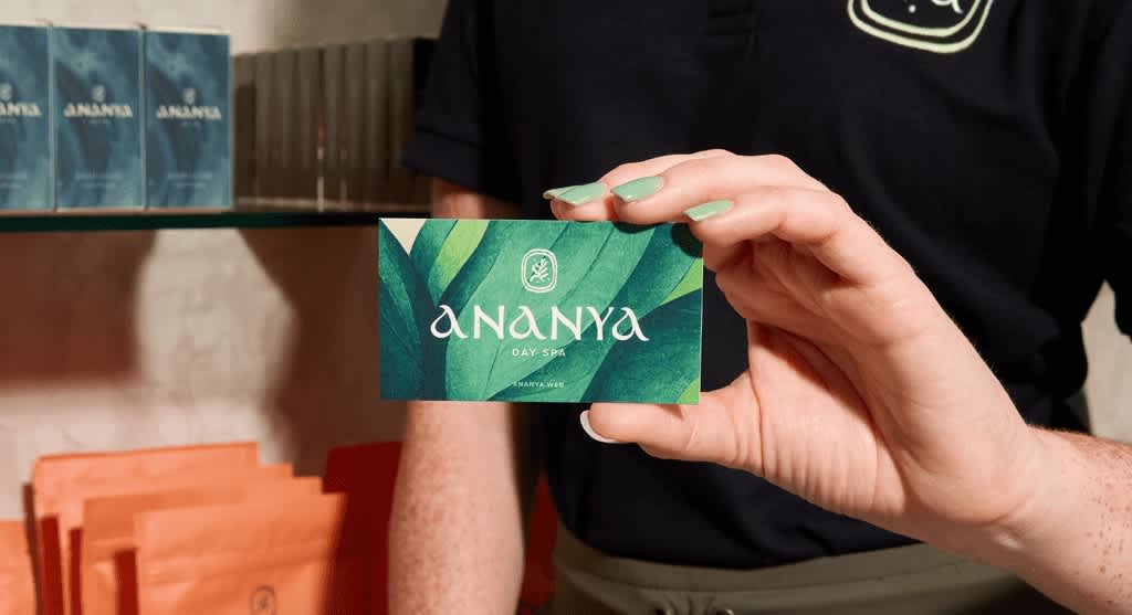 Business owner holding up business card with shades of green leaf design and a small cream logo and cream “Ananya” text.