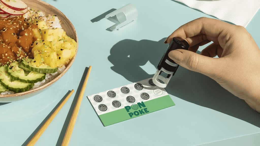 Green and white poke shop business card being stamped for a tenth time on a table next to a bowl of poke.