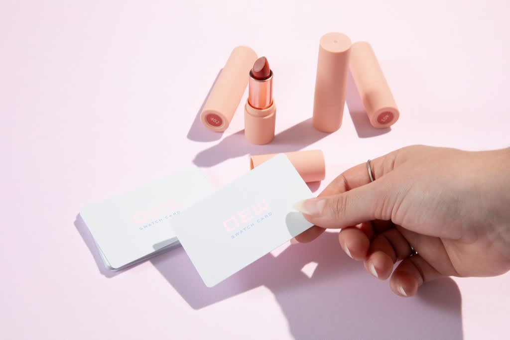 Hand holding up a simple white rounded edge business card with small pink and black text “Dew, Swatch Card” next to some lipsticks.