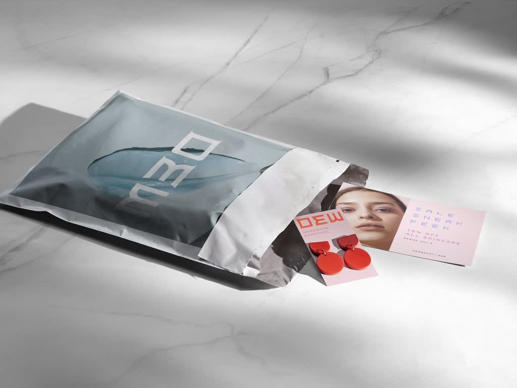 Silver DEW-branded mailer with a colorful promotional postcard and red handmade earrings displayed beside the open package.