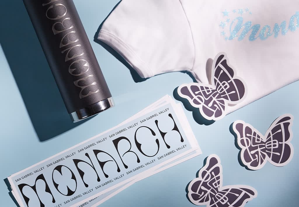 Custom butterfly-shaped stickers and logo stickers for Monarch San Gabriel Valley, displayed alongside a branded tumbler and white T-shirt.