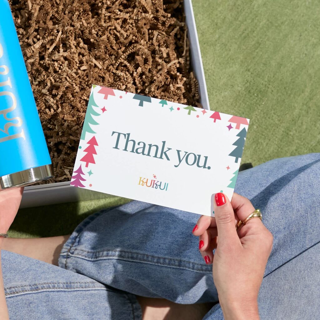 Colorful holiday-themed thank-you card held above an open gift box filled with crinkle-cut paper and a blue tumbler.